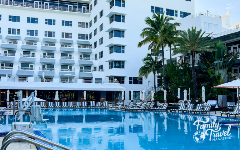 Hotel building with pool in front of it, surrounded by palm trees and chairs