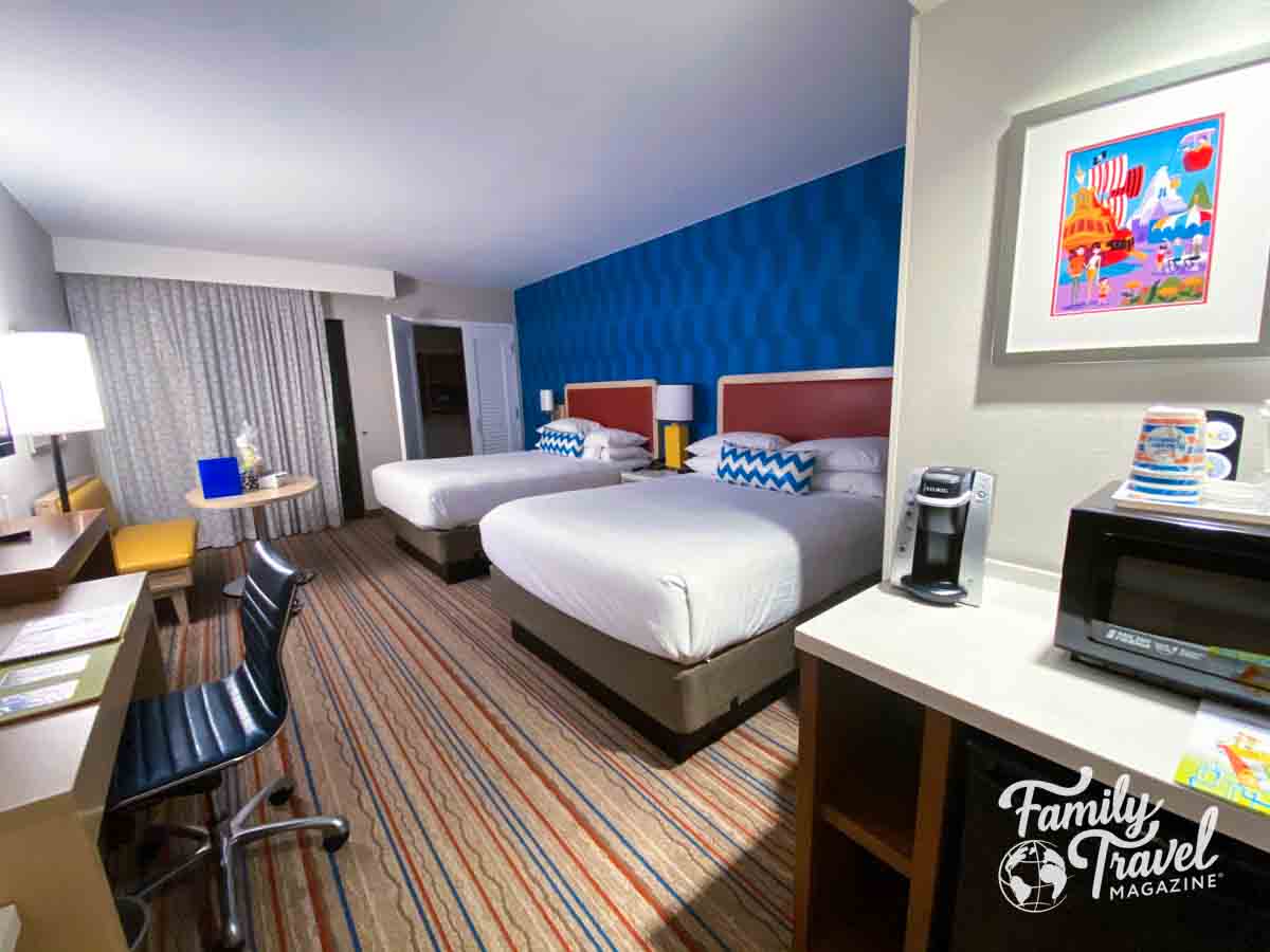 A hotel room with two queen beds, a desk, a seating area, a coffee maker, a microwave, and colorful decor