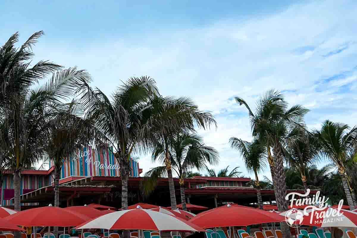 Slice of Paradise sign on building with umbrellas, palm trees, and beach chairs in foreground. 